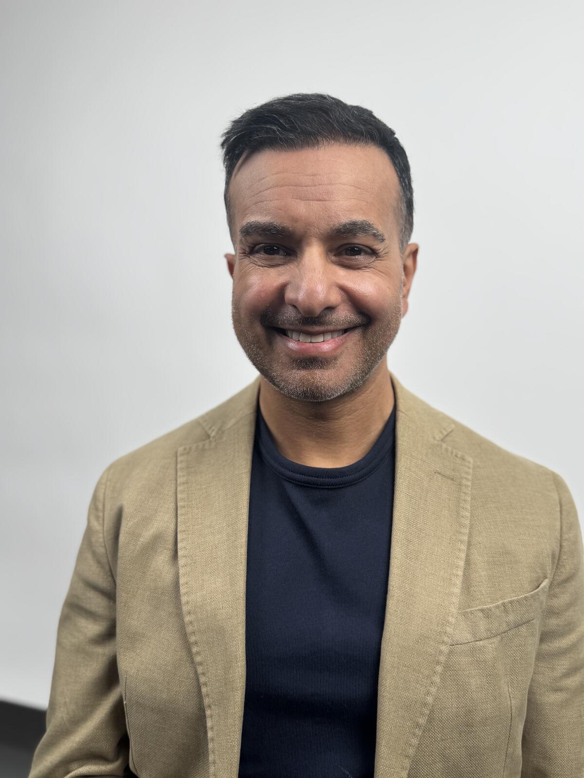 Sohail Ahmed, author, in a tan blazer over a navy crew-neck tee against a light gray background.